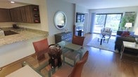This single-floor residence showcases a thoughtfully designed open-concept layout that seamlessly connects the dining and living areas. The dining space is anchored by a glass-top table paired with richly upholstered cognac leather chairs, creating an elegant yet inviting setting. A decorative oval mirror mounted above a lattice-front console table adds a stylish focal point to the dining area, while a granite-topped counter or bar is visible nearby, suggesting a practical transition toward the kitchen.

The living area extends naturally from the dining space, featuring warm hardwood flooring that runs throughout and a plush area rug grounding a contemporary seating arrangement. A glass-top coffee table with a sleek metal frame completes the sitting area. Large floor-to-ceiling windows flood the space with natural light and offer expansive views of lush greenery outside, enhancing the overall sense of openness. Built-in shelving with decorative accents adds character near the kitchen area. The cohesive design palette of neutral tones, warm wood finishes, and modern furnishings creates a refined, welcoming ambiance throughout the space.