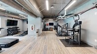 This fitness center at 2940 Falstaff Rd in Raleigh, NC offers a thoughtfully designed workout space with an industrial-chic aesthetic. The exposed concrete ceiling with visible ductwork and piping creates a raw, urban character, complemented by light wood-look flooring that adds warmth throughout the open floor plan. Linear LED lighting runs the length of the space, ensuring bright, even illumination across all workout zones.

The facility is comprehensively equipped with SportsArt cardio machines, including treadmills and ellipticals, positioned near a large window that provides natural light and outdoor views of the surrounding area. A dedicated strength training section features a multi-station cable machine, a full rack of dumbbells, and a weight bench, while additional cable and resistance machines line the perimeter. A wall-mounted flat-screen television is centrally positioned for entertainment during workouts.

The space is accented with framed artwork along the grey-painted walls, adding a polished touch to the utilitarian environment. A dual water fountain station near the entrance provides convenient hydration access. Rubber floor mats are strategically placed beneath equipment zones to protect both the flooring and users. The overall layout balances open movement space with dedicated equipment areas, making it a functional and well-appointed fitness facility.