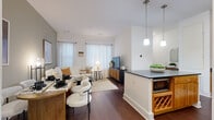 This well-appointed one-bedroom, two-bathroom residence at 80 Fawcett Street in Cambridge, MA offers a thoughtfully designed single-floor layout. The open-concept living and dining area is anchored by rich dark hardwood floors and filled with natural light from multiple windows dressed in sheer white curtains. A central kitchen island with a dark stone countertop and built-in wine rack doubles as a breakfast bar, seamlessly connecting the kitchen to the living space. The kitchen itself features warm wood cabinetry, dark stone countertops, stainless steel appliances including a refrigerator, range, and dishwasher, and pendant lighting overhead. The spacious primary bedroom includes a distinctive green accent wall, hardwood flooring, and natural light from a large window. A second bedroom offers a cozy layout with wall décor and natural light. Both bathrooms are finished with clean, modern details — one featuring a deep soaking tub set in white subway tile with brushed nickel fixtures, and the other with a dark wood vanity, white countertop, and light tile flooring. Throughout the unit, a neutral color palette and contemporary finishes create a cohesive, modern aesthetic.