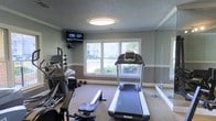 This single-floor fitness room offers a well-organized workout space designed to accommodate a variety of exercise needs. The room features a treadmill as a centerpiece, complemented by additional cardio equipment including an elliptical and a rowing machine, making it suitable for diverse training routines. A rack of dumbbells lines one wall, providing options for strength training exercises.

The space is finished with neutral gray walls accented by white crown molding and chair rail trim, lending a clean and polished appearance. A large, centrally placed flush-mount ceiling light ensures consistent illumination throughout the room. A wall-mounted flat-screen television adds entertainment value for users during workouts.

Natural light filters in through double windows along one wall, offering views of surrounding greenery and contributing to an open, airy feel. A full-length mirror along an adjacent wall enhances the sense of space while allowing users to monitor their form during exercise. The carpeted flooring adds a degree of comfort underfoot. Overall, the room provides a functional and well-appointed environment for fitness activities.