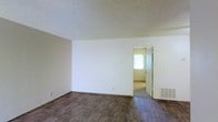 This single-floor space presents a straightforward, functional layout with a clean and neutral aesthetic throughout. The main living area features dark hardwood-style flooring that contrasts with the bright white walls, creating a simple yet balanced visual foundation. A popcorn-textured ceiling adds a classic finishing detail overhead.

The open wall space provides flexibility for furniture arrangement, while a floor vent indicates a built-in HVAC system for climate control. A doorway connects the main living area to an adjoining room, which receives natural light from a window visible through the opening, contributing a sense of brightness and continuity between spaces. The transition between the dark flooring in the main area and the lighter flooring in the adjacent room subtly defines each zone.

The overall ambiance is minimal and move-in ready, with neutral tones throughout that offer a versatile backdrop suited to a variety of interior styles. The uncluttered layout allows the architectural simplicity of the space to be immediately apparent, making it easy to envision how the rooms can be utilized.