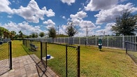 Located at 3640 Blue Fin Way in Fort Myers, Florida, this community dog park offers a spacious, fully enclosed outdoor area designed with pets and their owners in mind. The grounds are covered in lush green grass and lined with a dense hedge border, providing a natural and inviting atmosphere. Tall Florida palm trees are scattered throughout, offering shade and a distinctly tropical aesthetic that complements the sunny Southwest Florida climate.

The perimeter is secured by a black chain-link fence with a dedicated entry gate, ensuring pets can roam freely and safely. A bright blue pet bench is conveniently situated within the space, giving owners a comfortable place to sit and supervise their animals. Waste disposal stations are thoughtfully placed throughout the area, promoting cleanliness and ease of use. The 3D aerial view reveals the elongated layout of the park, with what appears to be a small agility or play feature toward one end, adding an interactive element to the space.

The backdrop features the light blue and white exterior of the surrounding residential buildings, framed by fluffy clouds and open sky. A street lamp provides illumination for evening visits. Overall, this dog park delivers a clean, functional, and welcoming outdoor amenity that reflects a well-kept residential community in the heart of Fort Myers.