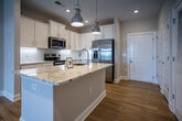 Located at 5060 City Station Dr in Collegedale, Tennessee, this single-story unit offers a well-designed, open-concept layout with modern finishes throughout. The kitchen serves as a central highlight, featuring white shaker-style cabinetry extending to the ceiling, a white subway tile backsplash, granite countertops, a central island with an undermount sink, and a full suite of stainless steel appliances including a refrigerator with ice maker, range, over-the-range microwave, and dishwasher. Polished chrome pendant lights hang above the island, complementing the recessed lighting overhead. Hardwood-style flooring flows through the kitchen and into the adjacent living area, which benefits from vaulted ceilings, a ceiling fan, and French doors that bring in natural light and outdoor views. An in-unit laundry area is conveniently located just off the kitchen. The bedrooms are carpeted and each include a ceiling fan and a walk-in closet with built-in rod storage. The bathrooms are finished with wood-look flooring, white vanities with granite countertops, and natural light from windows. The primary bathroom features a frameless glass sliding shower enclosure with chrome fixtures, adding a clean, contemporary touch to the overall aesthetic.