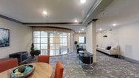 This single-floor space makes an immediate impression with its standout curved glass partition wall, which gracefully sweeps across a large portion of the interior and floods the space with natural light from the surrounding windows. The architectural design blends contemporary and classic elements, highlighted by recessed ceiling lighting, subtle tray ceiling detailing, and warm neutral wall tones accented by dark wood trim.

The layout is thoughtfully divided into distinct seating zones. One area features a round wooden table paired with vibrant orange chairs, offering a casual yet refined setting. Adjacent to this, a pair of sleek, curved black leather chairs with chrome frames surround a small round table, lending a sophisticated lounge feel. Along the far wall, a light-toned sofa with accent pillows provides additional comfortable seating beneath a modern pendant lamp.

Decorative potted topiary plants are positioned near the glass wall, adding a touch of greenery and elegance. The flooring throughout features a richly patterned carpet in muted, earthy tones that anchors the space. Structural columns are integrated seamlessly into the design. Overall, the space conveys a polished, professional ambiance suited for social or business gatherings.