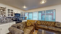 This single-floor space is thoughtfully designed to balance comfort and functionality. The open-concept living area is anchored by a generously sized tufted sectional sofa, complemented by warm wood-look flooring and a lower half-wall accent in a stone-textured finish that adds visual depth and character throughout. Large windows along one wall flood the living space with natural light and provide views of the surrounding outdoor area, creating a bright and welcoming atmosphere.

The kitchen is efficiently laid out and well-equipped, featuring stainless steel appliances including a refrigerator, dishwasher, and range with an over-the-range microwave. White cabinetry — including glass-front upper cabinets — pairs cleanly with granite-style countertops and a striking mosaic tile backsplash in earthy tones, giving the kitchen a modern, polished look. The layout connects seamlessly to the living area, making the space feel open and cohesive.

A separate office room offers a private, functional workspace equipped with a wall-mounted television, a desk setup with monitors, and access to the adjacent work area through elegant French doors with glass panels. The consistent use of crown molding, wood-look flooring, and textured stone wainscoting ties the entire space together with a unified, refined aesthetic. A glass-panel entry door provides a welcoming entrance with additional natural light.