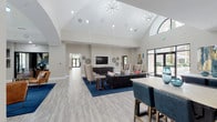 This single-story amenity space is defined by its dramatic architectural design, most notably the soaring vaulted ceiling that crowns the central lounge area. A sputnik-style chandelier hangs from the peak, complementing recessed lighting throughout and enhancing the airy, open atmosphere. The ceiling transitions into clerestory windows and arched transoms, drawing abundant natural light deep into the interior while framing views of lush outdoor greenery.

The main lounge is generously proportioned, featuring light wood-look tile flooring that runs throughout the space. Rich navy blue area rugs anchor distinct seating zones, including a welcoming entry lounge with cognac leather chairs and a larger gathering area furnished with a dark sectional sofa, accent chairs, and a wall-mounted television atop a sleek credenza. Decorative circular wall art adds visual texture to the neutral warm-beige walls.

A dedicated kitchenette and café-style bar area features two long marble-topped communal tables with teal upholstered stools, rich wood cabinetry, white upper cabinets, a beverage refrigerator, and pendant lighting with woven rattan shades. Full-height glass doors provide direct access to an outdoor patio, seamlessly connecting the interior to the exterior environment. The overall ambiance is modern, polished, and inviting.