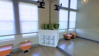 This single-floor fitness and wellness studio is thoughtfully designed to support a variety of exercise activities. The space features large windows fitted with roller shades that allow abundant natural light to filter in while maintaining privacy, creating a bright and energizing atmosphere. Light hardwood-style flooring runs throughout, contributing to a clean and modern aesthetic.

A central white storage unit serves as a functional focal point, providing open cubby shelving below and housing rolled green yoga or exercise mats on top, keeping equipment organized and easily accessible. Full-length mirrors line one wall, a standard feature in fitness spaces that allows users to monitor their form during workouts and visually expands the room. Ceiling-mounted TV bracket arms are positioned overhead, suggesting screens can be used for guided workouts or instructional content.

Orange swivel ottomans on chrome bases are placed around the perimeter, offering flexible seating options for rest or stretching. The overall design balances practicality and comfort, making the studio suitable for yoga, stretching, or light fitness activities. The neutral wall tones and coordinated furnishings give the space a cohesive, professional appearance.