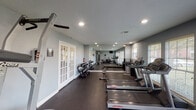 Located at 4720 Reading Rd in Rosenberg, TX, this dedicated fitness center offers a comprehensive workout environment within a single-floor layout. The space is finished with durable dark rubber flooring throughout, providing both safety and functionality for a wide range of exercises. Recessed lighting keeps the room well-lit, while multiple windows fitted with blinds allow natural light to filter in, creating an open and energizing atmosphere.

A standout feature is the large wall-length mirror that visually expands the space and allows users to monitor their form during workouts. The facility is equipped with Life Fitness treadmills, an elliptical trainer, a rowing machine, and a Hoist cable resistance machine, catering to both cardio and strength training needs. A full dumbbell rack offers a variety of free weight options, complemented by a weight bench and stability ball for versatile training.

A wall-mounted flat-screen TV provides entertainment during workouts, and a water dispenser ensures easy hydration access. White French doors with sidelights connect the space to the outdoors, enhancing ventilation and adding a welcoming architectural touch. The neutral gray walls and textured ceiling complete the clean, functional aesthetic of this well-appointed fitness facility.