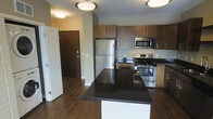 This single-floor residence showcases a thoughtfully designed open-concept kitchen that serves as the focal point of the living space. The kitchen is outfitted with a full suite of stainless steel appliances, including a french door refrigerator, a gas range with oven, and an over-the-range microwave. A central island with a striking dark granite countertop provides additional prep space and features built-in electrical outlets for added functionality. The cabinetry is finished in a warm medium-toned wood with flat-panel doors, offering ample storage. A tile backsplash adds a clean, contemporary detail behind the cooking area. Rich, dark hardwood flooring extends throughout the visible areas of the unit, lending warmth and cohesion to the space. Flush-mount ceiling light fixtures provide consistent illumination across the kitchen and entryway. The front entry, visible from the kitchen, features a solid dark wood door and is conveniently positioned near a closet, contributing to a practical and organized layout. The overall aesthetic is modern and polished.