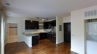 This single-floor space offers a clean, modern aesthetic anchored by an open-concept kitchen and living area. The kitchen is a standout feature, outfitted with bold dark cabinetry that contrasts beautifully against light granite countertops and a classic white tile backsplash. Stainless steel appliances, including a gas range, microwave, and refrigerator, provide a sleek, contemporary finish. A peninsula counter extends from the cabinetry, offering additional prep space and a natural divider between the kitchen and the main living area. Track lighting installed on the ceiling illuminates the kitchen efficiently, highlighting the space's functional layout. Rich, warm-toned hardwood flooring runs throughout, adding depth and continuity to the open floor plan. The walls are finished in a neutral, light tone that keeps the space feeling bright and airy. An electrical panel is neatly integrated into the wall near the entryway, and a dark-finished door anchors the entry point of the unit. The overall ambiance is modern and understated, blending practical design elements with contemporary style to create a cohesive and inviting living environment.