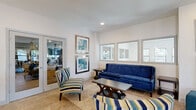 This well-appointed single-floor amenity space is thoughtfully designed for socializing and entertaining, offering a cohesive blend of modern style and functional layout throughout its distinct zones. The lounge area sets a welcoming tone with a tufted royal blue velvet sofa, coordinating striped accent chairs, teal ottomans, and a sleek coffee table resting on a woven area rug. Light tile flooring runs throughout, and framed abstract artwork in silver and gold frames adds a polished, curated touch to the bright white walls. A row of interior windows connects the lounge to adjacent spaces, enhancing openness and flow. The billiards room is a standout feature, centered around a full-size pool table with blue felt beneath a ceiling-mounted projector, flanked by large windows that invite natural light and garden views. Lush potted plants and a dark accent wall add depth and character to the space. The private dining area accommodates a large group around an extended table with upholstered chairs, a ceiling fan overhead, and multiple windows providing ample natural light. A decorative sideboard with ornate metalwork completes the room. The kitchen features a generous L-shaped white quartz island with seating for multiple guests, contemporary globe pendant lighting, stainless steel appliances, and a clean tile backsplash — ideal for catered events or communal use.