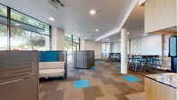 Located at 28850 Dixon St in Hayward, California, this single-floor commercial space offers a thoughtfully designed, contemporary environment ideal for collaboration and community gathering. The open-plan layout is anchored by a central structural column and features patterned carpet tiles in neutral tones accented with vivid turquoise inserts, adding a dynamic visual element throughout the floor.

The space is divided into several functional zones. A casual lounge area near the entrance incorporates modular privacy pods with teal and gray paneling, providing semi-enclosed seating for focused work or quiet conversation. Adjacent to this, floor-to-ceiling glazed windows flood the space with abundant natural light and offer unobstructed views of the tree-lined streetscape outside.

A dedicated dining or collaboration area toward the rear features high-top tables paired with modern bar-height chairs set against a decorative patterned accent wall, adding warmth and texture to the otherwise clean, modern aesthetic. A built-in kitchenette with dark cabinetry, a large-screen display, and stainless steel appliances serves the space with convenience.

A separate windowed alcove provides a quieter seating area with sleek pedestal chairs and a countertop workspace along the window wall, ideal for individual use. Recessed ceiling lighting throughout ensures consistent illumination across all zones, while glass-paneled entry doors maintain a sense of openness and connectivity with the surrounding building.