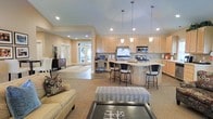 This single-story home showcases an open-concept layout that connects the living, kitchen, and dining areas into one cohesive, flowing space. The vaulted ceilings rise dramatically overhead, amplifying the sense of volume and openness throughout the main living area. Recessed lighting is strategically placed across the ceiling, complemented by a series of elegant pendant lights that hang above the kitchen island, adding both task lighting and visual interest.

The kitchen is well-appointed with light wood cabinetry, a stainless steel refrigerator, built-in microwave, and a gas range. The curved kitchen island provides additional seating with stylish bar-height stools, making it a natural gathering point. A tile backsplash adds a subtle decorative element to the cooking area.

Adjacent to the kitchen, a built-in desk area offers a functional workspace. The living area features neutral-toned flooring, a comfortable sofa with decorative pillows, and an upholstered bench, all centered around a dark-finished coffee table. Large glass doors at the rear of the space invite natural light and provide views to the outdoors, enhancing the home's bright, airy ambiance.