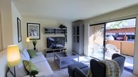 This single-floor residence offers a thoughtfully arranged and comfortable living environment. The living room serves as the heart of the space, featuring a light, neutral color palette with warm beige walls and carpeted flooring that contributes to a cozy, inviting atmosphere. Large sliding glass doors flood the room with abundant natural light and provide direct access to a private outdoor patio area, where seating is available for relaxing al fresco.

The living area is well-organized and functional, anchored by a plush sofa with green accent pillows and complemented by coordinating gray upholstered armchairs. A low-profile entertainment center houses a flat-screen television, flanked by open display shelving adorned with trailing green plants that add a refreshing natural touch. A matching gray display cabinet with glass-front doors provides additional storage and display space. A glass-top coffee table centered in the seating arrangement ties the room together with a clean, contemporary aesthetic.

Wall art and potted indoor plants enhance the space's character, creating a lived-in yet tidy ambiance. The patio visible through the sliding doors suggests the space extends naturally to an outdoor seating area, further expanding the overall sense of openness and light throughout this single-level home.