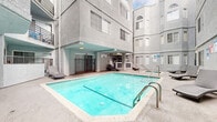 Located at 5555 Harold Way in the vibrant Hollywood neighborhood of Los Angeles, this shared amenity space serves as a central gathering hub within a multi-story residential building. The space is defined by its inviting outdoor pool area, which is the clear centerpiece of the complex. The generously sized swimming pool, filled with crystal-clear turquoise water, is complemented by a separate hexagonal spa, offering residents a relaxing retreat. Multiple lounge chairs are positioned poolside, providing comfortable spaces for sunbathing and leisure. The pool deck is enclosed on multiple sides by the building's stucco-clad exterior, creating a semi-private, courtyard-like atmosphere. A covered patio area adjacent to the pool provides shaded seating and connects to a secured entry point with wrought iron gate details. The interior entrance corridor features large-format tile flooring, textured stucco walls, recessed lighting, and a ceiling fan, creating a cool and functional passageway. Glass doors at the end of the corridor offer a clear sightline to the pool area beyond, reinforcing the seamless indoor-outdoor connection that defines this amenity space.