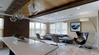 This single-floor space masterfully combines rustic charm with contemporary sophistication. The living area immediately draws the eye upward to its striking ceiling, where rich, dark exposed wooden beams contrast beautifully against a warm wood-paneled ceiling. Recessed lighting complements the abundant natural light that pours in through a series of large windows dressed in sheer, neutral-toned curtains, creating a bright and airy atmosphere throughout.

A prominent brick column adds an industrial, textural element that anchors the space and bridges its rustic and modern sensibilities. The living area is thoughtfully furnished with a sleek sectional sofa in cool grey, paired with modern swivel accent chairs on polished chrome bases, all arranged to encourage comfortable conversation and relaxation.

Pendant light fixtures with handblown glass shades hang gracefully overhead, serving as both functional lighting and sculptural focal points. The flooring transitions seamlessly from a high-gloss light stone surface near the kitchen into warm wood-toned planks in the living zone, subtly defining each area.

A large piece of abstract wall art provides a bold visual accent, while a tray ceiling detail in the living area adds architectural depth. The overall ambiance is one of refined comfort — warm yet polished, open yet intimate.