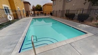 This community recreational area is centered around a generously sized swimming pool with a clean, tiled interior and stainless steel entry handrails, offering a refreshing outdoor amenity for residents. The pool deck is finished in smooth concrete and is enclosed by a sturdy black iron perimeter fence, ensuring safety and privacy throughout the space.

A standout architectural feature is the covered cabana structure, which frames the pool area with elegant arched openings supported by rounded stucco columns. Recessed lighting in the cabana ceiling provides illumination for extended use, while the open arches maintain unobstructed sightlines to the pool. The stucco finish on the columns and surrounding walls is consistent with the warm, neutral tones of the adjacent multi-story residential buildings visible in the background.

Access to the pool area is controlled through a decorative wrought iron gate, featuring ornamental scrollwork at the top arch, adding a classic aesthetic touch to the functional security element. Conveniently located within the covered cabana area are dedicated restroom facilities, with clearly marked separate entrances for men and women. The louvered doors on the restroom entries allow for ventilation while maintaining privacy. Overall, the space is designed for practical, communal outdoor recreation with thoughtful architectural detailing throughout.
