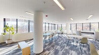 This contemporary single-floor space is designed to foster collaboration and informal gathering, combining open-plan functionality with a clean, modern aesthetic. The layout is divided into distinct yet fluid zones, including a lounge area and multiple dining and breakout seating clusters, all unified by stylish patterned carpet tiles in muted blue and grey tones.

The lounge area features a sleek black leather sofa, contemporary white swivel chairs, and a mustard yellow ottoman, arranged to encourage relaxed conversation. Large windows along the perimeter flood this zone with natural light and offer pleasant views of surrounding trees and the urban streetscape.

The dining and collaborative zones are furnished with oval and round tables in blue and white finishes, paired with modern shell-style chairs in white, cream, and sage green. Floor-to-ceiling windows wrap around the corner of the space, maximizing daylight and providing expansive outdoor views. A prominent structural column anchors the center of the room without impeding the open feel.

Overhead, recessed linear LED lighting complements the natural light throughout the day. Glass-paneled doors at the entry provide visibility into adjacent areas, reinforcing the open and connected character of the space. Potted plants add a touch of greenery, enhancing the overall welcoming and professional ambiance.