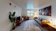This single-floor unit offers a thoughtfully organized layout with multiple distinct living spaces connected by light wood-style flooring throughout. The living room is a welcoming, open area bathed in natural light from a large window with garden views, complemented by smooth white ceilings and clean white walls that enhance the sense of space. It comfortably accommodates a full seating arrangement and entertainment setup, with greenery adding a fresh, lively touch.

The kitchen is efficiently designed with warm wood cabinetry extending to the upper walls, granite-style countertops, a built-in electric range, and a stainless steel sink — all positioned alongside a window that keeps the space bright and airy.

The unit features multiple bedrooms, each with its own character. One room offers dual windows with serene outdoor views, hardwood-style flooring, and ample space for a full bed and desk area. Another bedroom is a cozy, well-lit space with a window, wood armoire, and room for a sitting area. A third bedroom provides a quieter atmosphere with white-painted block walls and warm accent lighting. A fourth bedroom features two windows and flexible furniture placement.

The full bathroom is clean and practical, featuring tile flooring, white cabinetry with overhead storage, grab bar accessibility features, and a shower with a curtain enclosure.