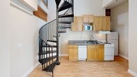 Located at 2018-2030 University Ave in Berkeley, CA, this two-floor loft offers a cleverly designed layout that maximizes its compact footprint. The main floor serves as the heart of the space, featuring a combined kitchen and dining area equipped with light maple cabinetry, granite countertops, a full suite of appliances including a dishwasher and microwave, and a stainless steel sink. Light hardwood flooring runs throughout, complementing the warm, neutral tones of the walls. A standout architectural feature is the sleek black metal spiral staircase, which serves as both a functional and visual centerpiece connecting the two floors. Soaring ceilings with exposed wood beam accents add character and a sense of openness. A bright window allows natural light to pour in, further enhancing the airy feel. The main floor also includes a full bathroom, finished with marble-style tile flooring, a white vanity, and a tub/shower combination with sliding glass doors. Upstairs, the loft level provides a versatile living or sleeping area with gray laminate flooring, a mirrored sliding-door closet, and a skylight that fills the space with additional natural light. The upper level overlooks the main floor, reinforcing the open, loft-style character of the home.