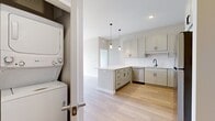 This efficiently designed one-bedroom, one-bathroom unit offers a thoughtfully organized layout across a single floor. The open-concept kitchen and living area serves as the heart of the unit, where light wood-tone flooring extends throughout, creating a warm, cohesive aesthetic. The kitchen features a quartz waterfall island with an undermount sink and matte black faucet, white shaker cabinetry, stainless steel appliances including a refrigerator, stove, and microwave, and pendant lighting that adds a modern touch. The living area benefits from generous natural light and direct access to a private balcony through a floor-to-ceiling sliding glass door with vertical blinds, offering open outdoor views.

The bedroom is carpeted for comfort, finished in neutral tones, and includes a dedicated walk-in closet equipped with wire shelving and hanging rods for organized storage. The full bathroom features a white shaker vanity with a clean white countertop, matte black fixtures, a large mirror, and a shower-tub combination. A separate in-unit laundry area houses a stacked washer and dryer alongside wire shelving for additional storage. Clean white walls, consistent trim, and a neutral palette give the entire unit a bright, modern ambiance.