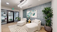 Located at 1133 Harding Place in Charlotte's vibrant 28204 corridor, this single-floor space masterfully blends wellness and professional functionality with a polished, contemporary aesthetic. The reception and lounge area immediately sets an elevated tone, anchored by a textured blue-grey mosaic accent wall adorned with a large-scale abstract painting in teal and white. Two plush, curved lounge chairs with ottomans rest atop a geometric patterned rug beneath a sculptural wave-form light fixture, creating a calm and inviting atmosphere. Light wood herringbone flooring runs throughout, lending warmth and visual continuity to the entire space. Lush tropical plants are strategically placed to reinforce the spa-like ambiance. A dedicated treatment room offers a professional massage table positioned beneath recessed lighting, complemented by floor-to-ceiling sage velvet drapes, white subway tile, warm wood cabinetry, and a framed mirror — delivering a refined, clinical-yet-comfortable environment. A glass-partitioned meeting room enclosed by sleek black-framed doors provides a visually open yet acoustically private workspace, visible from the lounge and equipped for collaborative use. Together, these spaces form a cohesive, design-forward suite ideal for wellness or professional services.