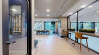 Located at 550 Westlake Park Blvd in Houston, Texas, this thoughtfully designed single-floor lounge space offers a contemporary and inviting environment well-suited for collaboration, casual meetings, or relaxed work sessions. The open layout is anchored by a generously sized living area that seamlessly blends multiple seating configurations to accommodate a variety of needs.

The lounge features a long, built-in blue banquette with colorful accent pillows paired with round pedestal tables and an assortment of accent chairs, creating a comfortable and social atmosphere. A floor-to-ceiling dark wood open shelving unit serves as a striking architectural divider within the space, adding visual depth and a sophisticated design element.

A sleek bar-height counter runs along a wall of large glass windows, lined with warm-toned leather stools — an ideal setting for individual work or casual conversation. The expansive glazing throughout floods the interior with natural light and offers views of lush greenery outside, enhancing the connection to the outdoors.

Light wood-look flooring anchors the space, complemented by a multi-toned geometric area rug in the seating zone. Recessed ceiling lighting and wall-mounted sconces provide layered illumination throughout. A large display screen mounted on a wood-paneled wall adds a modern, functional touch. The overall ambiance is polished, vibrant, and professionally inviting.