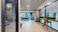 Located at 550 Westlake Park Blvd in Houston, Texas, this thoughtfully designed single-floor lounge space offers a contemporary and inviting environment well-suited for collaboration, casual meetings, or relaxed work sessions. The open layout is anchored by a generously sized living area that seamlessly blends multiple seating configurations to accommodate a variety of needs.

The lounge features a long, built-in blue banquette with colorful accent pillows paired with round pedestal tables and an assortment of accent chairs, creating a comfortable and social atmosphere. A floor-to-ceiling dark wood open shelving unit serves as a striking architectural divider within the space, adding visual depth and a sophisticated design element.

A sleek bar-height counter runs along a wall of large glass windows, lined with warm-toned leather stools — an ideal setting for individual work or casual conversation. The expansive glazing throughout floods the interior with natural light and offers views of lush greenery outside, enhancing the connection to the outdoors.

Light wood-look flooring anchors the space, complemented by a multi-toned geometric area rug in the seating zone. Recessed ceiling lighting and wall-mounted sconces provide layered illumination throughout. A large display screen mounted on a wood-paneled wall adds a modern, functional touch. The overall ambiance is polished, vibrant, and professionally inviting.
