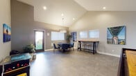 This single-floor community clubhouse is thoughtfully designed to accommodate a variety of social and recreational activities. The space is characterized by soaring vaulted ceilings that lend an open, airy feel throughout, while a consistent palette of warm neutral tones is punctuated by bold teal accent walls that energize each zone.

Upon entry, a welcoming corridor leads into the main gathering area, where a large communal table with mixed seating — including teal upholstered chairs — sits atop a decorative area rug beneath a striking geometric pendant light. Landscape and nature photography displayed as large canvas prints add a Pacific Northwest character to the walls.

Adjacent to this central space is a dedicated game and entertainment zone featuring a shuffleboard table, wall-mounted flat-screen television, and a classic arcade cabinet with surrounding stools — ideal for casual recreation. A separate card room hosts a full-sized poker table encircled by upholstered seating beneath a warm drum pendant fixture.

A secondary dining and meeting area offers a round table arrangement with additional bar-height seating along a wall-mounted counter, bathed in natural light from large windows overlooking green outdoor surroundings. Herringbone-patterned tile and wood-look flooring flow throughout, tying the various functional areas together into a cohesive, well-appointed communal retreat.