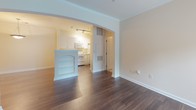 Located at 6450 Mercantile Dr E in Frederick, Maryland, this single-floor residence offers a well-organized layout with a clean, neutral aesthetic throughout. The living area features rich, dark hardwood-style flooring and flows seamlessly into the kitchen through a distinctive arched entryway accented with crown molding. A charming built-in bookshelf unit frames the transition between spaces, adding character to the open feel. The kitchen is outfitted with white cabinetry, a full suite of white appliances including a coil-top range, over-the-range microwave, and refrigerator, all complemented by light countertops and track lighting overhead. The bedrooms are carpeted in a soft, neutral tone and receive generous natural light through large windows with blinds overlooking greenery outside. One bedroom features a ceiling fan for added comfort. The primary bathroom includes a full tub-shower combination with a glass enclosure, a white vanity, and wood-look flooring. A second bathroom offers a white vanity with ample cabinet storage, a large mirror, and matching hardwood-style flooring. The overall palette of warm beige walls, white trim, and contrasting dark floors creates a cohesive and inviting atmosphere throughout this thoughtfully designed space.