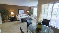 This single-floor residence offers an open-concept layout that seamlessly connects the living and dining areas, creating a cohesive and functional living environment. The living area features plush carpeted flooring, warm taupe-toned accent walls, and white upper walls and ceiling that brighten the space. A comfortable sofa adorned with decorative pillows anchors the seating area, complemented by a cushioned armchair and a dark rectangular ottoman that doubles as a coffee table. Framed wall art and a floor lamp add to the room's polished aesthetic, while a tall indoor plant introduces a natural element near the window.

The dining area, positioned in the foreground, features a round glass-top table surrounded by dark brown upholstered chairs, offering an elegant contrast to the softer tones of the living space. A floral centerpiece provides a refined decorative touch. Natural light flows generously through multiple windows fitted with horizontal blinds, illuminating the open layout throughout. The overall ambiance is warm, contemporary, and welcoming, with a cohesive neutral color palette that ties both functional areas together harmoniously.