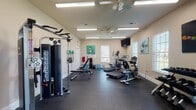 This fitness center is a comprehensively outfitted single-floor exercise space designed to support a full range of workout routines. The room features durable rubber flooring throughout, providing both safety and comfort underfoot during exercise. Overhead, flush-mounted fluorescent lighting combined with ceiling fans ensures the space remains well-lit and well-ventilated during use.

The cardio section is equipped with multiple treadmills, an elliptical trainer, and a stationary upright bicycle, offering users a variety of aerobic exercise options. Strength training is supported by a multi-function cable machine with a pull-up bar attachment, an adjustable weight bench, and a full rack of dumbbells spanning a wide weight range. Additional selectorized strength machines are positioned along the perimeter of the room.

Multiple windows fitted with white blinds line the walls, allowing natural light to filter into the space while maintaining privacy. A wall-mounted flat-screen television provides entertainment during workouts. Water coolers and several large water jugs are available for hydration. The neutral beige walls are accented by a collection of bold, colorful athletic-themed canvas paintings, adding an energetic and motivating ambiance to the overall environment.