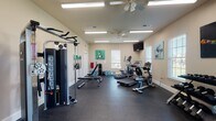 This fitness center is a comprehensively outfitted single-floor exercise space designed to support a full range of workout routines. The room features durable rubber flooring throughout, providing both safety and comfort underfoot during exercise. Overhead, flush-mounted fluorescent lighting combined with ceiling fans ensures the space remains well-lit and well-ventilated during use.

The cardio section is equipped with multiple treadmills, an elliptical trainer, and a stationary upright bicycle, offering users a variety of aerobic exercise options. Strength training is supported by a multi-function cable machine with a pull-up bar attachment, an adjustable weight bench, and a full rack of dumbbells spanning a wide weight range. Additional selectorized strength machines are positioned along the perimeter of the room.

Multiple windows fitted with white blinds line the walls, allowing natural light to filter into the space while maintaining privacy. A wall-mounted flat-screen television provides entertainment during workouts. Water coolers and several large water jugs are available for hydration. The neutral beige walls are accented by a collection of bold, colorful athletic-themed canvas paintings, adding an energetic and motivating ambiance to the overall environment.