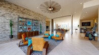 This contemporary single-floor clubhouse is designed to offer a dynamic and inviting communal environment. The entrance foyer greets visitors with large floor-to-ceiling glass panels and expansive windows that flood the space with natural light, while offering clear views of the surrounding greenery outside.

The main lounge area is anchored by a striking exposed brick accent wall adorned with open metal shelving displaying curated décor. Bold, mid-century-inspired seating in warm amber tones is complemented by turquoise accent pieces, all arranged atop layered area rugs over large-format tile flooring. An oversized decorative ceiling fan serves as a focal point overhead.

A generous open corridor lined with colorful framed artwork and wall-mounted plants connects the lounge to additional seating zones, including a casual dining area outfitted with mixed-material tables and chairs. Floor-to-ceiling windows here continue to blur the boundary between interior and exterior.

The fully equipped kitchen features rich dark wood cabinetry, a hexagonal mosaic tile backsplash, stainless steel appliances, and a white-topped island with bar seating — ideal for community gatherings and events.

A game room with a billiards table, co-working stations with task seating, and additional lounge nooks round out this multifunctional space, making it a well-appointed hub for both productivity and leisure.