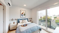 This single-floor space showcases a clean, contemporary design with a cohesive neutral palette of crisp whites and warm wood-toned flooring that runs throughout. The bedroom serves as the centerpiece of the unit, offering generous natural light through large sliding glass doors that open onto a private balcony, providing pleasant outdoor views of the surrounding neighborhood. The room comfortably accommodates a full-sized bed and includes built-in wall art placement and matching nightstands, giving it a well-appointed, balanced feel. A full-length black-framed mirror is positioned in the hallway area adjacent to the bedroom, enhancing the sense of depth and light in the transitional space. The bathroom features warm hardwood-style flooring, a clean white toilet, a mounted towel bar, and a decorative wall display of hanging planters with greenery, adding a fresh, organic touch to the otherwise minimalist design. Throughout the space, the architectural detailing remains consistent — smooth white walls, recessed lighting, and clean baseboards contribute to the modern aesthetic. The sliding glass door in the bedroom frames views of neighboring structures and tree canopy, connecting the interior to its urban surroundings. Overall, the space presents a bright, functional, and stylish living environment with a well-considered layout and modern finishes.