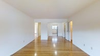 This single-floor space offers a clean, minimalist aesthetic defined by polished hardwood floors that reflect abundant natural light, creating a warm and inviting atmosphere throughout. The main living area is generously proportioned, with smooth white walls and a neutral palette that lends a bright, airy feel. The open layout connects seamlessly to adjoining spaces, with multiple doorways leading to additional rooms, enhancing the sense of flow and connectivity. Visible from the main area, the kitchen features a stainless steel refrigerator and receives natural light from a window, contributing to an open and functional culinary space. A baseboard heating unit is visible near the kitchen entry, indicating a practical heating system integrated throughout. Additional doorways on the right side of the space suggest access to further rooms, potentially including storage or utility areas. The consistent hardwood flooring and crisp white walls carry throughout, unifying the space with a cohesive and timeless design. Overall, the space presents a straightforward, well-lit layout ideal for comfortable everyday living.