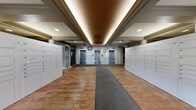 This building lobby presents a polished and functional entryway designed to serve a high-occupancy residential building. The space is divided into two connected areas that flow seamlessly into one another. The primary entry zone features warm terracotta brick-style tile flooring accented by a central runner mat, flanked on both sides by extensive banks of white parcel and mailbox lockers — providing convenient package management for residents. Three stainless steel elevator doors are prominently positioned along the rear wall, accompanied by digital display panels and a secure access keypad. The ceiling in this zone features a recessed tray design with warm indirect uplighting and recessed downlights, creating an inviting and well-lit atmosphere. Decorative crown molding adds a refined architectural touch throughout. The adjacent lounge area transitions to plush carpeting and showcases elegant coffered ceilings supported by fluted columns. Framed artwork lines the neutral gray walls, contributing a cultured ambiance. A generous floor-to-ceiling glass window wall at the far end bathes this area in natural light and offers views of mature outdoor trees and landscaping. Raised planter beds with lush tropical foliage further enhance the biophilic character of the space, creating a welcoming and serene lobby environment.