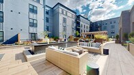 Located at 347 E Fifth Ave in Columbus, Ohio's vibrant Short North area, this thoughtfully designed outdoor amenity space offers a dynamic and versatile environment for relaxation and recreation. The courtyard is framed by a modern multi-story building clad in bold blue-gray siding with private balconies, creating an enclosed and intimate atmosphere open to the sky.

The space is organized into several distinct zones. Multiple lounge areas feature generously cushioned wicker and upholstered seating arrangements set atop light wood-toned pavers, ideal for gathering and socializing. A wooden pergola serves as a central architectural focal point, providing partial shade and a defined gathering space beneath. A sleek linear water feature anchors one section of the courtyard, adding a calming ambient element.

A well-maintained putting green offers a recreational outlet, complete with flags and additional casual seating nearby. Raised wooden planters filled with ornamental grasses, flowering plants, and small trees are distributed throughout, adding greenery and natural texture to the hardscape. A separate enclosed turf area, bordered by black iron fencing, provides open lawn space. Additional patio sections with container plantings and café-style seating offer quieter retreats. The entire space benefits from abundant natural light and open sky views, creating a welcoming outdoor destination year-round.