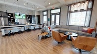 This single-floor space is a well-appointed communal lounge designed for both relaxation and social gatherings. The open-concept layout seamlessly integrates a kitchen bar area with a comfortable living space, creating a cohesive and inviting environment.

The kitchen bar area features dark cabinetry, light-colored countertops, and industrial-style bar stools, providing a casual setting for dining or socializing. A wall-mounted television and stainless steel appliances enhance the functionality of this zone, while a textured stone backsplash adds visual depth.

The lounge area is anchored by a generous sectional sofa in neutral gray, complemented by a cognac leather accent chair and an organic live-edge coffee table. The eclectic furniture arrangement encourages conversation and relaxation. Wood-look tile flooring extends throughout the space, adding warmth while remaining easy to maintain.

Tall windows fitted with dark-framed transoms flood the interior with natural light, offering views of the surrounding exterior. High ceilings with recessed lighting and pendant fixtures contribute to the spacious, airy feel. The overall design blends industrial and contemporary elements for a polished, modern ambiance.