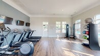 Located at 1980 W Main St in Central, South Carolina, this well-appointed fitness center offers a clean, bright, and welcoming environment for exercise. The single-floor space is thoughtfully designed with light gray walls, crown molding, and warm hardwood-style flooring that runs throughout, lending a polished yet functional aesthetic.

The main workout area is generously open and filled with natural light streaming through multiple large windows, several of which offer views of an outdoor pool area. A glass-panel door provides direct access to the exterior. Recessed lighting supplements the natural brightness, ensuring a well-lit environment at any hour.

The exercise room is equipped with a variety of cardio machines, including treadmills and an elliptical, as well as a multi-station cable strength machine, a weight bench, and a dumbbell rack stocked with free weights and medicine balls. A wall-mounted flat-screen television is positioned near the cardio equipment for entertainment during workouts.

A compact kitchenette area is integrated into the space, featuring white cabinetry, granite countertops, a stainless steel refrigerator, and a sink — providing convenient access to refreshments. The overall ambiance is bright, modern, and inviting, making this fitness center a practical and comfortable amenity.