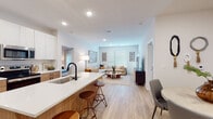 This well-appointed single-floor residence showcases a thoughtfully designed open-concept layout that seamlessly connects the kitchen, dining, and living areas. The kitchen is a standout feature, equipped with stainless steel appliances, a large island with a white quartz countertop and bar seating, a matte black faucet, and a two-tone cabinet design pairing white upper cabinets with warm wood-toned lower cabinets. A dedicated in-unit laundry room with a full-size washer and dryer is conveniently accessible from the kitchen.

The open living area is bright and airy, bathed in natural light from large windows, and features light wood-look flooring throughout. A dedicated dining nook provides an intimate space for meals. The two bedrooms are carpeted for comfort, each featuring ceiling fans and generous windows with blinds and drapes. The primary bedroom offers ample space, while both rooms maintain a calm, neutral aesthetic.

The bathrooms echo the kitchen's design language, with wood-grain vanity cabinetry, white quartz countertops, large mirrors, and light flooring. The primary bathroom includes a spacious walk-in shower and a long double-sink vanity. Clean white walls throughout lend the residence a bright, contemporary feel.