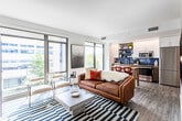 This single-floor residence showcases a thoughtfully designed open-concept layout that seamlessly connects the living, dining, and kitchen areas. The living room is a standout space, lined with expansive floor-to-ceiling windows on two sides that flood the interior with natural light and frame dynamic urban views of surrounding buildings and tree-lined streets. The room is anchored by a cognac leather sofa, a concrete coffee table, and a bold navy-striped area rug, complemented by a gallery wall of framed artwork that adds a curated, artistic character. Light hardwood-style flooring runs throughout, contributing to a cohesive and contemporary aesthetic.

The kitchen features warm wood cabinetry, a neutral countertop, a stainless steel sink, a built-in dishwasher, and eye-catching blue subway tile backsplash. Open shelving provides practical display storage, while white upper cabinets maximize the sense of brightness. A compact dining area with red barstools connects the kitchen to the main living space.

The bathroom is clean and modern, with dark slate tile flooring, a wood-toned vanity with open shelving, a white quartz countertop, a large mirror, and a full-size soaking tub with herringbone tile surround. The overall ambiance throughout is urban, contemporary, and refined.