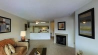 This single-story residence showcases an open-concept design that seamlessly connects the living and kitchen areas. The living room is anchored by a classic fireplace with a clean white surround, serving as a natural focal point of the space. Neutral-toned carpeting extends throughout the main living area, contributing to a warm and inviting atmosphere. A ceiling fan is centrally positioned to provide both comfort and lighting to the shared living space.

The kitchen is accessible via a pass-through counter, which enhances the open flow between the two areas while maintaining a defined cooking zone. The kitchen features white appliances, including a refrigerator and range, alongside upper cabinetry and under-cabinet lighting. A textured popcorn ceiling adds a traditional character throughout the space.

The walls are finished in soft neutral tones, providing a versatile backdrop for décor, as evidenced by the framed artwork displayed above the fireplace. The overall ambiance is casual and comfortable, with a practical layout that balances open living with distinct functional zones.