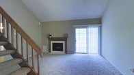 This two-story residence offers a thoughtfully arranged living space that balances comfort and functionality. The main living area serves as the heart of the home, centered around a classically framed fireplace with a warm wood mantel and white surround, providing a focal point that adds both character and coziness to the room. Soft, neutral-toned carpeting extends throughout the living area, contributing to a comfortable and inviting atmosphere. A large sliding glass door fitted with vertical blinds allows an abundance of natural light to fill the room while offering convenient access to the outdoor area beyond. The walls are finished in a warm, neutral tone that complements the overall aesthetic. A staircase with wooden railing is positioned along one side of the living area, providing access to the upper floor and adding a sense of vertical dimension to the space. The ceiling is smooth and well-proportioned, reinforcing the open and airy feel of the room. Overhead ventilation is present, ensuring year-round climate control. The overall ambiance is clean, understated, and move-in ready.