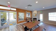 This single-story space offers a welcoming, contemporary atmosphere characterized by a thoughtful blend of warm and neutral tones. The most striking design element is a bold amber-orange accent wall that anchors the room, creating a vibrant focal point against the surrounding light gray walls. Recessed lighting throughout the ceiling provides consistent, well-distributed illumination, complementing the natural light that filters in through multiple windows fitted with horizontal blinds.

A central wooden dining table with sleek, modern chairs serves as the primary gathering feature, suitable for both casual meetings and shared meals. Nearby, a bar-height table with tall chairs offers an alternative seating arrangement, adding versatility to the layout. A gas fireplace set into the gray wall provides a cozy ambiance, accented by a decorative sunburst mirror mounted above it. The flooring features wood-look planks that add warmth and continuity throughout the space. A glass-panel door at the far end allows additional natural light to enter, while a secondary doorway connects to an adjacent area, enhancing the overall sense of openness and flow.