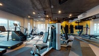 Located at 3883 S Moody Ave in Portland, Oregon, this well-appointed fitness center delivers a comprehensive workout environment within a thoughtfully designed single-floor space. The industrial-modern aesthetic is defined by exposed concrete ceilings with visible ductwork and structural columns, recessed lighting, and a bold geometric-patterned carpet that adds visual energy to the floor.

The main workout area is generously equipped with a full rack of dumbbells and kettlebells, multiple cable and resistance machines, and a dedicated stretching and functional training zone. A vibrant yellow accent wall featuring an energetic fitness mural anchors the free weights section, complemented by a wall-mounted flat-screen TV for entertainment during workouts. Floor-to-ceiling mirrors along one wall enhance the sense of space and allow for form monitoring.

A dedicated cardio zone houses treadmills and elliptical machines positioned directly in front of large windows, offering views of the surrounding Portland landscape and filling the space with natural light. Near the entrance, built-in wood shelving provides convenient storage, and a small refreshment station with a beverage cooler and cabinetry adds a practical amenity. The overall ambiance is motivating and modern, balancing industrial design elements with functional, high-quality fitness equipment.