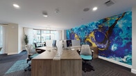 This single-floor professional space blends contemporary design with curated artistic expression, creating an environment that is both functional and visually inspiring. The most striking feature is a large-scale abstract mural spanning an entire wall in deep blues, teals, and gold — a dramatic focal point that anchors the open-plan workspace. Modern workstations with clean wood-toned desks and ergonomic mesh chairs are arranged to encourage productivity, while floor-to-ceiling windows along one side of the room flood the area with natural light and provide views of an outdoor courtyard and pool area. A dedicated dining or conference area features a sleek rectangular table with dark modern chairs, positioned directly beside the expansive glazed windows and illuminated by a contemporary linear pendant fixture. The lounge area offers a relaxed atmosphere with a curated arrangement of modern accent chairs, a round coffee table, and a textured driftwood sculptural installation that adds an organic, artistic counterpoint to the contemporary surroundings. Gallery-style artwork, including a large impressionistic landscape painting and several ornate gold convex mirrors, lines the neutral-toned walls, contributing to an upscale, gallery-like ambiance. Charcoal striped carpet tiles throughout unify the space while adding visual texture underfoot.