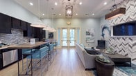 Located at 6880 Rivers Ave in North Charleston, SC, this single-floor community clubroom offers a well-appointed gathering space designed for both comfort and functionality. The interior is finished with light wood-look flooring and soft sage-green walls, creating a calm, welcoming atmosphere throughout.

The open layout seamlessly connects a fully equipped kitchen area to a comfortable lounge. The kitchen features dark espresso cabinetry, a mosaic tile backsplash, stainless steel appliances including a refrigerator and microwave, and a long reclaimed-wood communal table with teal upholstered barstools positioned beneath a trio of oversized pendant lights — ideal for casual dining or socializing.

The lounge area is anchored by a striking herringbone-patterned accent wall housing a mounted flat-screen television, complemented by a plush neutral sectional, a rustic wood-and-metal coffee table, and contemporary artwork in muted blue and green tones. A geometric lantern pendant light adds an elegant touch overhead.

French doors with glass sidelights flood the space with natural light and provide views of the surrounding community. The overall design balances modern and transitional aesthetics, making this a versatile and inviting communal space.
