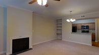 This single-floor residence offers a comfortable and functional layout designed for everyday living. The main living area serves as the heart of the home, centered around a classic fireplace that adds warmth and character to the space. Crown molding runs along the ceiling throughout, lending a refined, traditional touch to the interior. Soft, neutral-toned carpeting underfoot contributes to the room's inviting atmosphere, while the light beige walls keep the space feeling bright and open. A ceiling fan provides practical comfort, complementing the elegant chandelier that hangs nearby, offering layered lighting options. Built-in shelving along one wall provides convenient display and storage space, adding both functionality and visual interest. A pass-through window opens directly into the kitchen, where wood cabinetry and a dark refrigerator are visible, hinting at a well-appointed culinary space beyond. The overall ambiance is warm and classic, with thoughtful architectural details — including the crown molding and built-ins — that give the home a polished, timeless character well-suited for comfortable day-to-day living.
