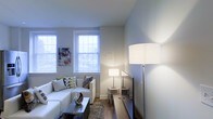 This two-floor space is designed with a clean, contemporary aesthetic that emphasizes comfort and functionality. The living area showcases a neutral color palette dominated by soft whites and cool grays, creating a calm and inviting atmosphere. Two large double-hung windows dressed with horizontal blinds allow an abundance of natural light to fill the room, illuminating the space and offering views of the surrounding greenery outside.

The living area is furnished with a white sofa accented by patterned throw pillows in muted greens and grays, complemented by a sleek, rectangular coffee table with a metallic finish. A plush area rug anchors the seating arrangement, adding warmth and texture to the hardwood flooring beneath. Layered lighting is a notable feature, with a tall floor lamp and coordinating table lamps providing ambient illumination throughout the room.

A staircase with a simple metal railing is visible, connecting the two floors and reinforcing the space's modern design sensibility. The walls are kept minimal, with a single piece of colorful artwork providing a subtle focal point. Overall, the space balances understated elegance with practical, livable design.